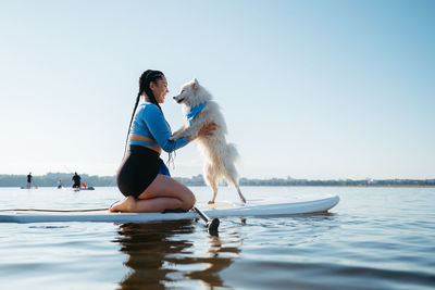 Happy young woman with locs hugging with her dog japanese spitz while sitting on sup board on lake