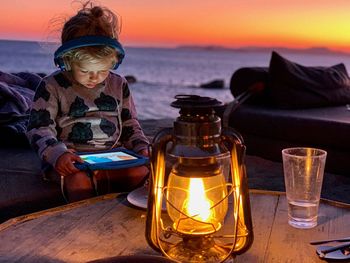 Boy using digital tablet by illuminated old-fashioned lantern at beach during sunset