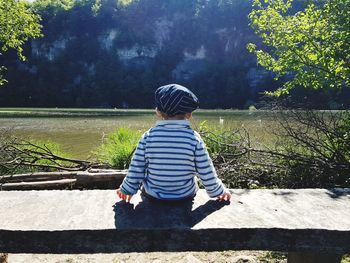 Rear view of boy sitting by lake on sunny day