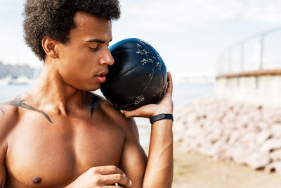 Shirtless young man exercising with fitness ball while standing at beach against sky