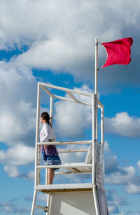 Young woman watches the water from a life guard tower on the beach, while a red flag flies 