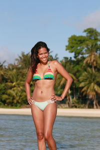 Happy young woman in bikini standing at beach against sky