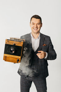 Portrait of young man holding camera while standing against white background