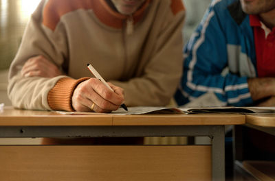 Midsection of man writing on paper while sitting by friend