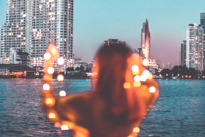 Rear view of woman photographing illuminated buildings by sea against sky