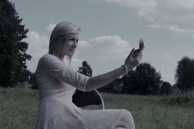 Side view of young woman holding hand mirror while sitting on chair at grassy field