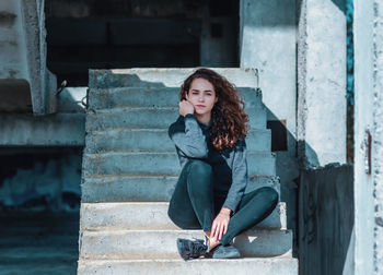 Charming girl with curly hair is sitting on stairs. crossed feet, hand near face.