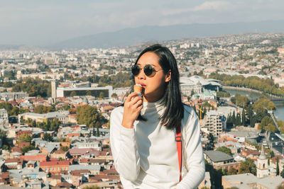 Woman eating ice cream while standing against buildings in city