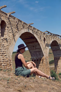 Woman traveler explorer in a black hat sit next to the destroyed old stone bridge in the summer