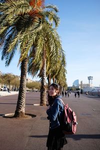 Portrait of woman standing by palm tree in city against sky