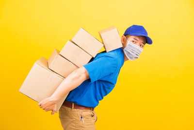 Side view of delivery person wearing mask holding cardboard box standing against yellow background