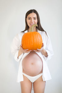 Portrait of smiling pregnant woman holding pumpkin against white background