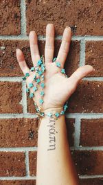 Cropped image of tattooed hand holding rosary beads against brick wall