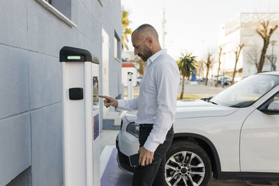 Man using automat standing by car at electric charging station