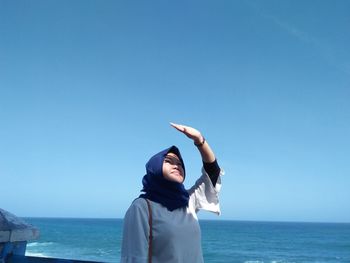 Woman shielding eyes while standing at beach against clear blue sky