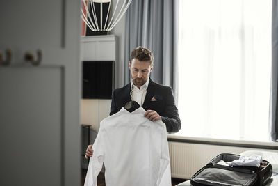 Businessman holding white shirt in coathanger against window at hotel room