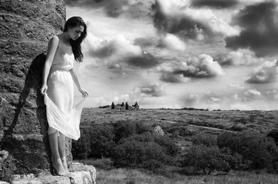 Full length of young woman standing by wall against cloudy sky