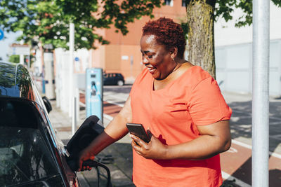 Happy woman holding mobile phone charging electric car on sunny day