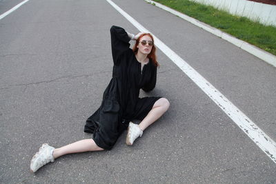 Fashion portrait of ginger woman in loose black dress and sneakers sitting posing on road outside 