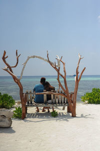 Rear view of couple sitting on bench at beach against clear sky