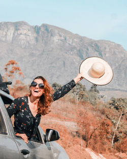Woman with hat in hand with body getting out of the car with serra do caraça in the background.