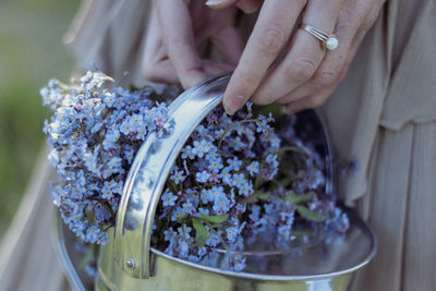 Close-up of a woman's hands holding a silver watering containing small blue and purple flowers.