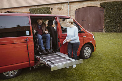 Senior woman holding remote control while helping man with disability in motorized wheelchair to disembark from van