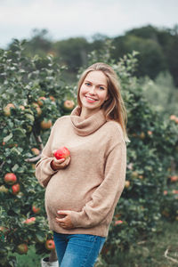 Portrait of smiling pregnant woman holding apple while standing by basket and trees