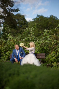 Bridal couple sitting on bench against plants at park