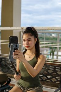 Portrait of young woman photographing while sitting on railing