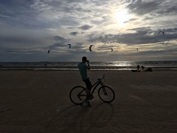 Man on bicycle at beach against sky during sunset