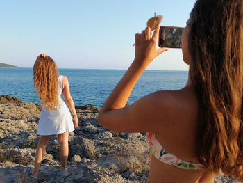 Two young girls make photos on the rocks with phone. wonderful landscape, blue sea and sky