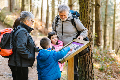 Grandparents hikers with kids reading information on wooden board while standing on path in woods in autumn during trekking