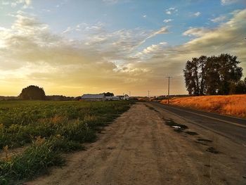 Dirt road amidst field against sky during sunset