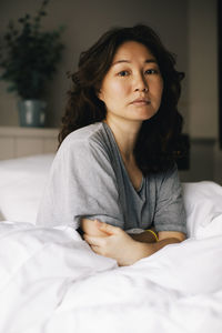 Portrait of lonely woman sitting on bed with white blanket at home