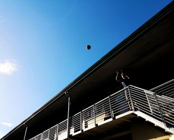 Low angle view of man throwing ball from building balcony against blue sky