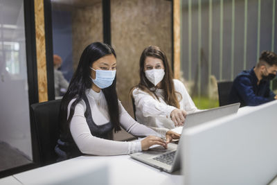 Multiracial female colleagues wearing masks sitting at table with laptop and discussing business project while working in coworking space