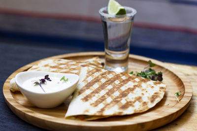 The glass of tequila with lime and portion of quesadilla on round wooden board, mexican cuisine