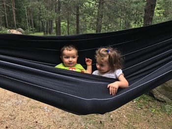 Siblings playing in hammock against trees at forest