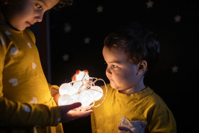 Side view of curious siblings in pajamas standing in dark room with illuminated garland