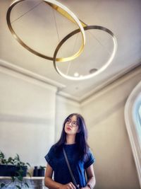 Asian woman in eyeglasses standing under circular led lights in white room.