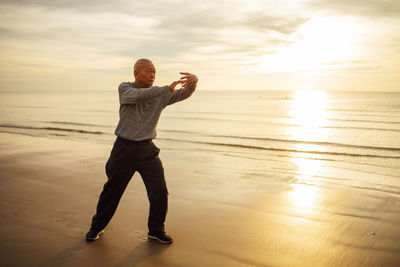 Senior man exercising while standing at beach against sky during sunset