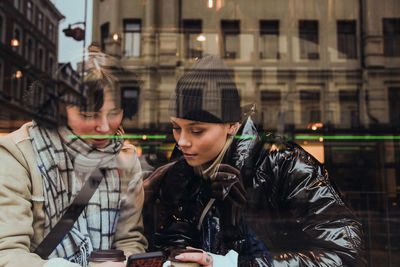 Young woman wearing warm clothing while looking at female friend using mobile phone in cafe seen through glass window
