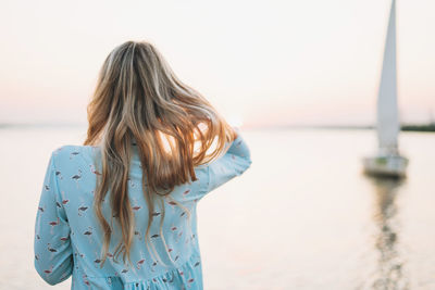 Beautiful blonde young woman in blue dress on pier on background of sailboat on sunset
