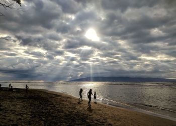 Silhouette children on shore at beach against cloudy sky during sunset