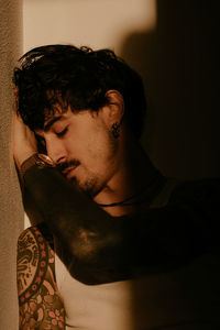 Young stylish guy with mustache and tattoos wearing white tank top and leaning against wall with back with closed eyes