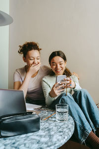 Happy young woman sharing smart phone with female teenage friend at home