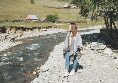 Brunette young woman traveler in poncho near mountain river, country side