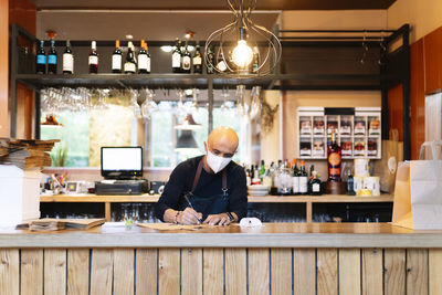 Mature male owner writing while working on bar counter during pandemic