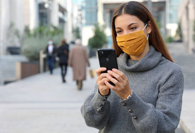 Young woman wearing mask using mobile phone while standing outdoors